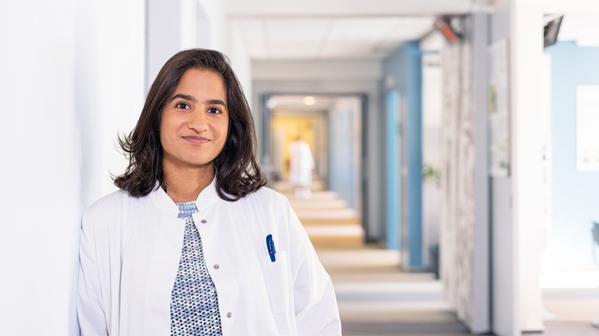 Eine junge Frau in einem weißen Kittel steht in einem Krankenhausflur und lächelt in die Kamera.